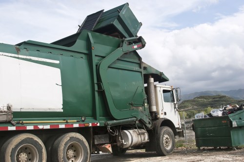 A large commercial waste collection truck situated outdoors on a gravel surface under a cloudy sky, with a backdrop of rolling hills and distant greenery. The truck features a white cab at the front with visible side mirrors and a black bumper. Its main body, designed for rubbish collection, is painted green and fitted with a hydraulic lifting mechanism that indicates it is in the process of loading or unloading waste. This mechanism includes a large, rectangular, metal hopper with a slightly weathered finish, positioned at the rear of the truck, which is open, revealing an empty interior. Adjacent to the truck on the right side, there is a green waste bin filled with assorted rubbish, including plastic bags and miscellaneous debris, placed on the gravel ground. The scene suggests a routine rubbish removal operation, typical of commercial waste services provided by companies such as Commercial Waste Removal Raynes Park, operating within the postal district associated with Raynes Park, South West London.