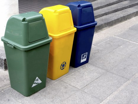 Three plastic rubbish bins are positioned on a paved pedestrian walkway, with the green bin on the left, the yellow bin in the middle, and the blue bin on the right. The green bin, made of smooth, matte plastic, features a rounded lid and a triangular recycling symbol on the lower front panel. The yellow bin has a rounded top and displays a small circular icon with arrows, indicating recycling, while the blue bin, also with a smooth surface and rounded lid, has a recycling symbol sticker on the lower front area. These bins are arranged in a line, aligned parallel to a building facade with steps leading up to the entrance, neighboring a modern, grey, textured paving surface typical of commercial and pedestrian areas within Raynes Park. The environment is an outdoor urban setting, suitable for waste disposal and rubbish collection services, with natural daylight illuminating the scene. Commercial Waste Removal Raynes Park would manage waste collection from such containers in local commercial and residential areas within the area code, offering professional rubbish removal tailored to local needs.