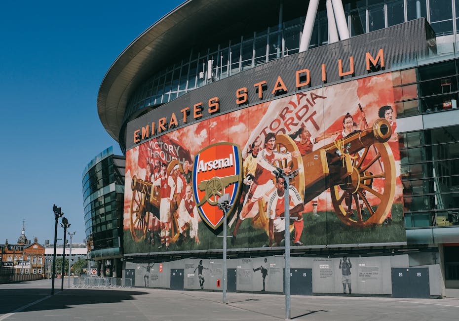 A large, modern stadium features a prominent curved exterior with glass panels and metallic finishes, displaying a vibrant mural on its facade that depicts historical soldiers operating cannons and ca