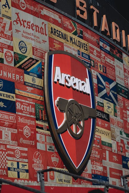 A large, modern stadium features a prominent curved exterior with glass panels and metallic finishes, displaying a vibrant mural on its facade that depicts historical soldiers operating cannons and ca