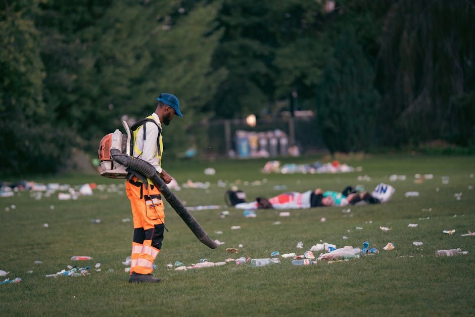 A waste management worker dressed in a high-visibility orange and black uniform, including reflective strips, is using a leaf blower or similar equipment to clear litter from a grassy outdoor area. Th