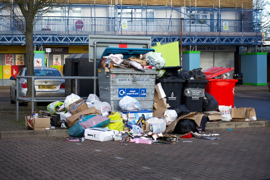A waste management worker dressed in a high-visibility orange and black uniform, including reflective strips, is using a leaf blower or similar equipment to clear litter from a grassy outdoor area. Th